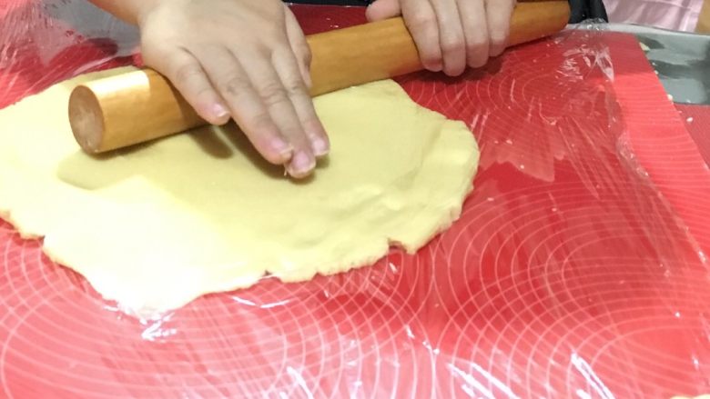 如何做出酥脆的卡通饼干,将面团放在案板上摁扁，在上面铺上一层保鲜膜，用擀面杖擀成0.3cm厚度的面饼，打开保鲜膜，开始用模具制作