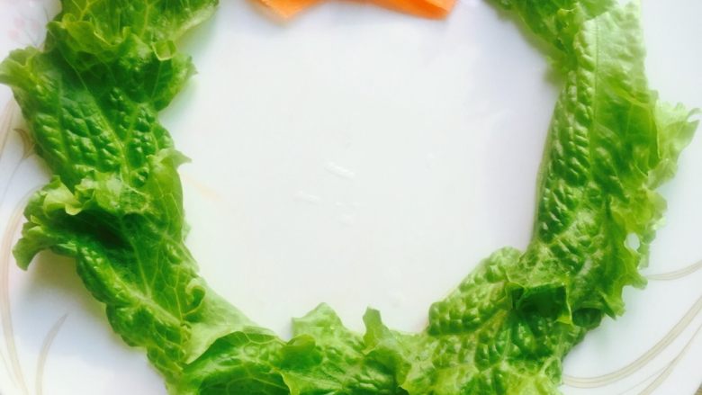 健康版圣诞蔬菜花环沙拉#乐享双节#,取一个白色盘子，将生菜撕成片沿着盘子的边缘摆成环状。取胡萝卜片打成蝴蝶结🎀