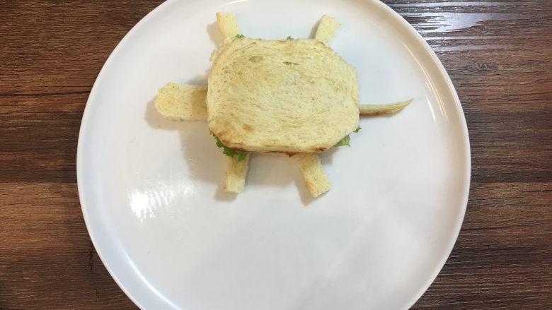 童趣早餐——乌龟全麦三明治,用切剩下的面包边角料切出乌龟的头、脚和尾巴，摆放在乌龟的身体旁边。