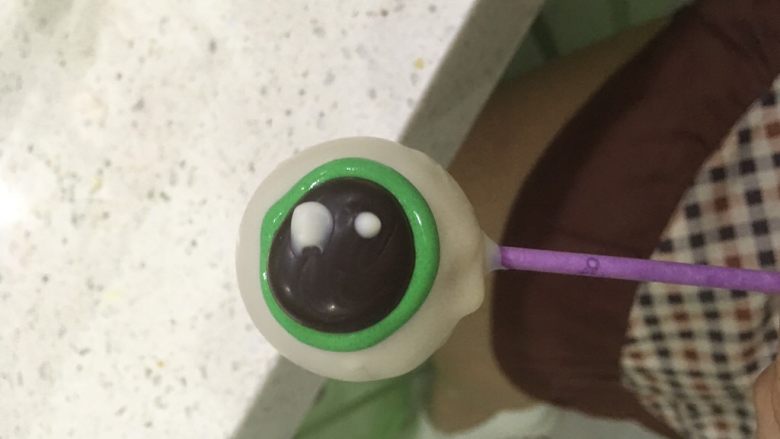 万圣节系列—眼球蛋糕棒,先用绿色糖霜话一个圆，然后把巧克力眼珠放到圆的中心位置，如图。