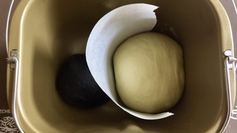 奶牛纹吐司（淡奶油版）,把两个面团中间隔上一张烘焙纸，避免混色，放入面包桶，盖上保鲜膜放在温暖处进行基础发酵。参考温度28°，时间约一个小时。