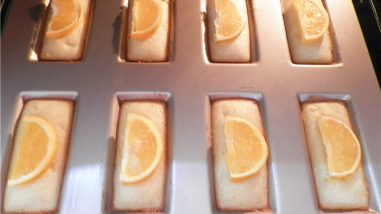 橙香费南雪,烤7分钟后，将橙子片分别码在：每个“金砖”的中间。该模具导热性很好， 再烤7分钟左右表面金黄即可。