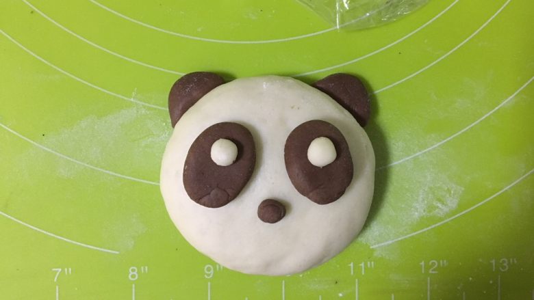 呆萌熊猫豆沙包,另取两个可可小面团，搓成椭圆形，按扁，沾一点水沾在熊猫的脸上，白面团粘在眼袋上，再取一点点可可面团作为鼻子