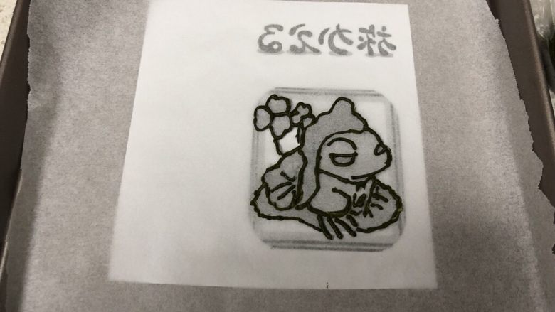 旅行青蛙蛋糕卷,把打印好的镜像的旅行青蛙图案上放上一张油纸，用最深色的抹茶黄油霜勾勒出轮廓