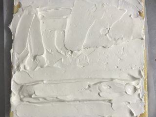 爱心草莓蛋糕卷,蛋糕片下垫上一张较大的油纸，将奶油抹在蛋糕片上，靠近身边的蛋糕卷用奶油堆一个小山丘。