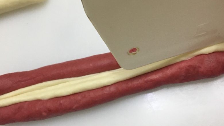 爱心💗吐司,把两个15克的白色面团搓成22厘米的棒状，把一根白色棒状面团摆入2条细的红色棒状面团之间，用刮板按压，使其固定。