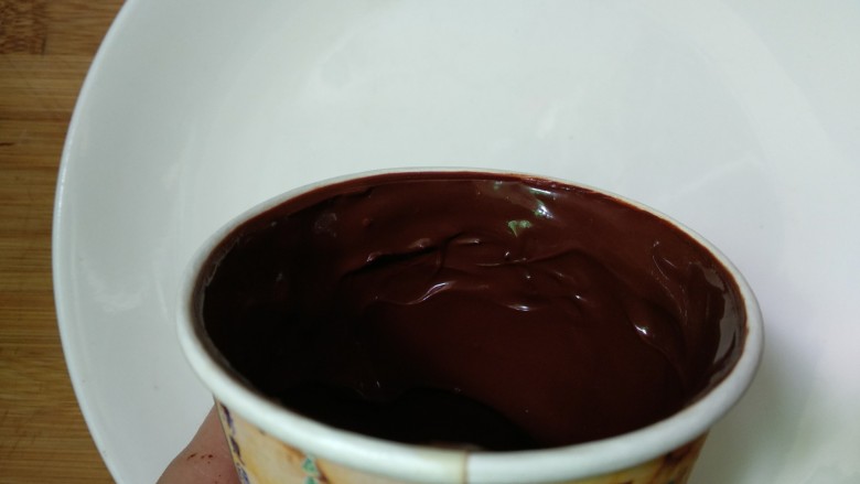 仿真早餐（芒果布丁+巧克力杯子）,一次性纸杯里倒入适量巧克力，快速转动杯子，让巧克力酱粘在杯壁上，放入冰箱冷藏15分钟。取出重新倒入巧克力酱，借助勺子把杯壁涂厚点，杯底巧克力也要厚，放入冰箱冷藏3小时（具体时间根据杯子大小调整）在油纸上画出杯把放入冰箱冷藏。