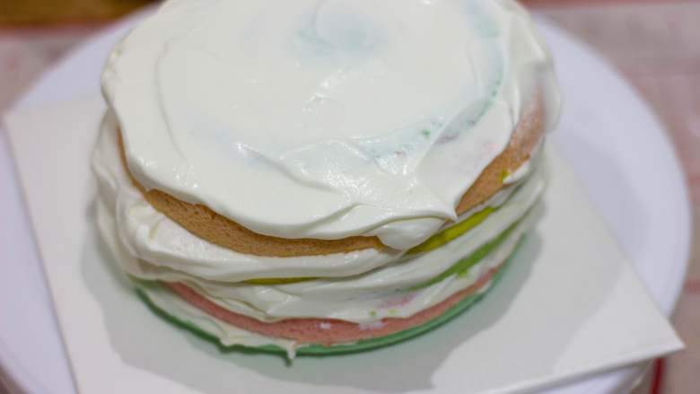 棋格奶油蛋糕，给生活画出一道彩虹,转动蛋糕台，让表面抹的更加均匀；再放一片彩色蛋糕胚，抹奶油，直到最后一片蛋糕胚；