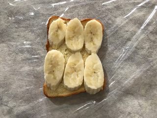 香蕉鸡蛋芝士三明治,在保鲜膜的中间位置放上一片吐司，并且均匀码上切成斜片的香蕉