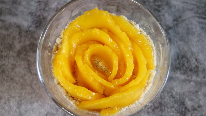 芒果木糠杯,最后在饼干屑的上面摆上芒花型,一片片的叠加摆出玫瑰花形状。