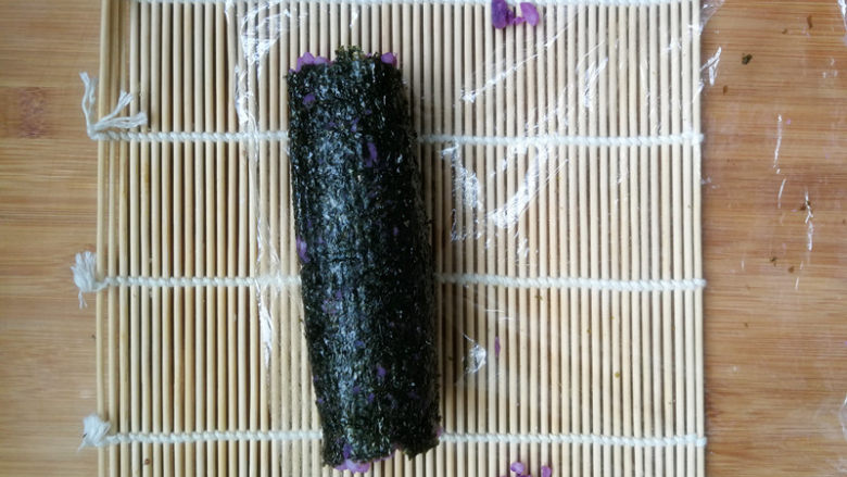 紫薯米饭香肠卷,从一头将寿司帘卷起来，卷紧，松开，挪动一下保鲜膜，继续往前卷，卷紧定型。