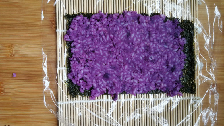 紫薯米饭香肠卷,寿司帘上铺上一层保鲜膜，放上一张大半张海苔。海苔上均匀地铺上紫薯米饭，用力压实。