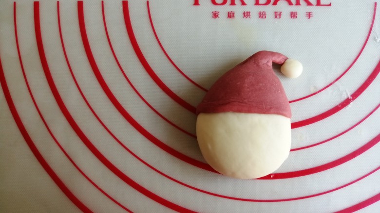圣诞老人馒头,将擀好的红色面团包着头部上方，捏成帽子形状，揪出一丁点备用的白色面团，放在帽尖上
