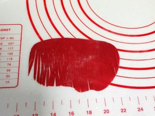 小福猪中国结馒头,取4克红色面团揉匀排气，擀成半椭圆状，用刮刀切细条状，顶部预留0.8厘米不要切断，之后从中间一切为二。