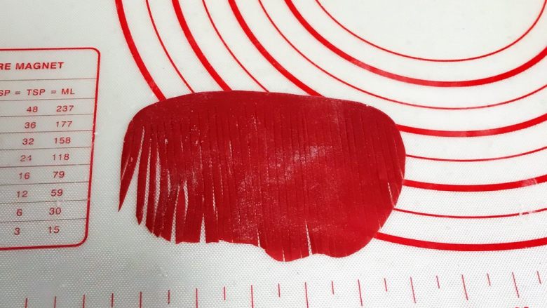 小福猪中国结馒头,取4克红色面团揉匀排气，擀成半椭圆状，用刮刀切细条状，顶部预留0.8厘米不要切断，之后从中间一切为二。