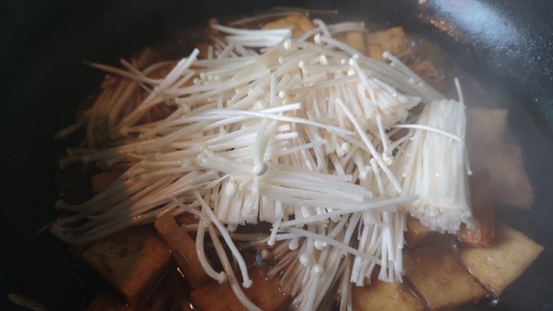 金针菇豆腐煲,锅里留少量油，放入蒜末炒香后倒入炸好的豆腐，翻炒两下倒入料汁，将金针菇放入锅里，加水炖至金针菇变色。