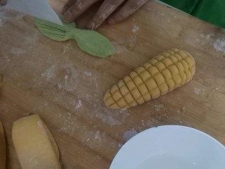 彩蔬玉米馒头,取一个绿色剂子用手搓成长条状，再压成叶子状，用刮刀压出纹路