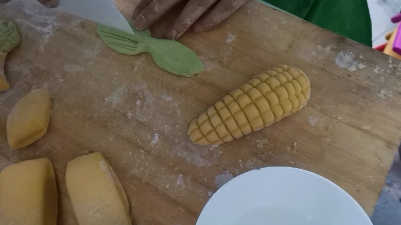 彩蔬玉米馒头,取一个绿色剂子用手搓成长条状，再压成叶子状，用刮刀压出纹路