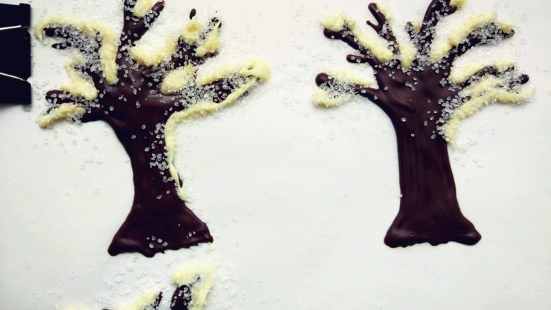 雪国的树——椰蓉巧克力杯蛋糕,静置片刻凝固后，用隔水融化的白巧克力，在树枝上画出积雪的样子，待白巧克力八成凝固时，撒上少许砂糖，静置备用