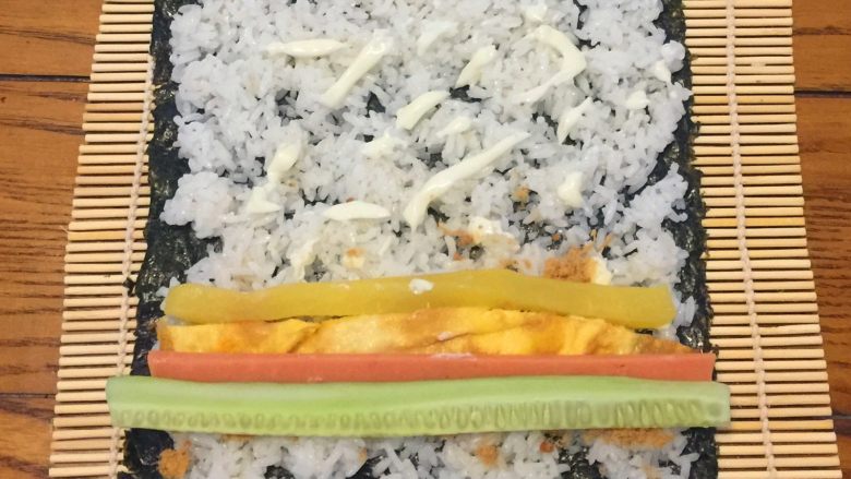 超简单寿司做法,把黄瓜，火腿肠，鸡蛋，调味萝卜平铺在肉松上，从自己这边往外卷