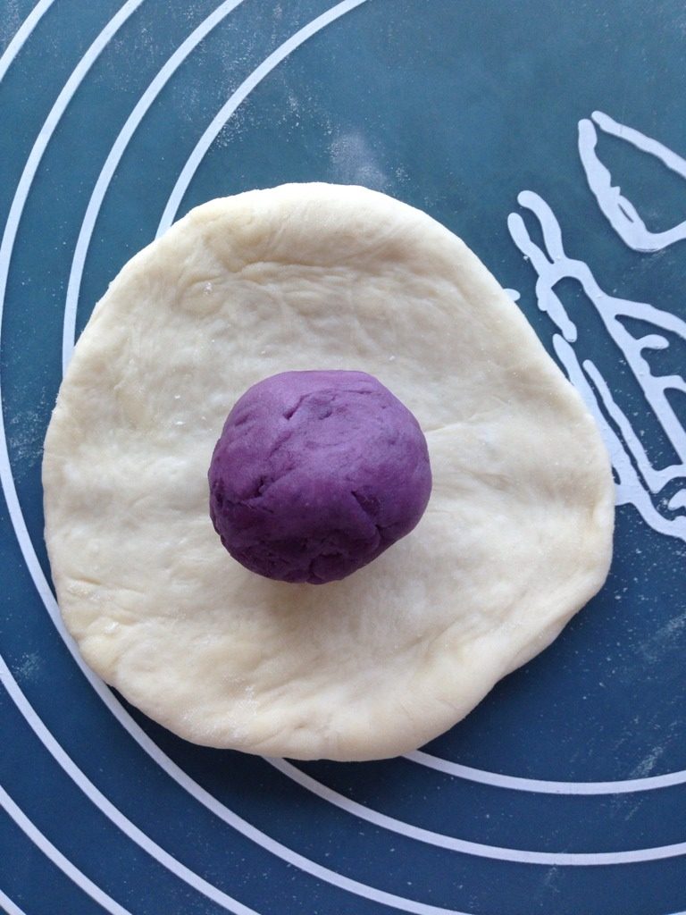 Q弹紫薯馅花朵面包,排好气后取出面团，分割成5等份，滚圆，放松10分钟。紫薯馅也分成5等份，搓圆。然后将小面团擀成圆形，翻面，包入紫薯馅。捏紧收口。